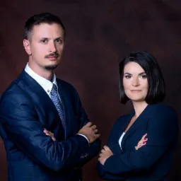 Portret dwojga prawników z Rzeszowa, mężczyzna w niebieskiej kraciastej marynarce i kobieta w granatowym garniturze, oboje z założonymi rękami na tle ciemnobrązowego tła.