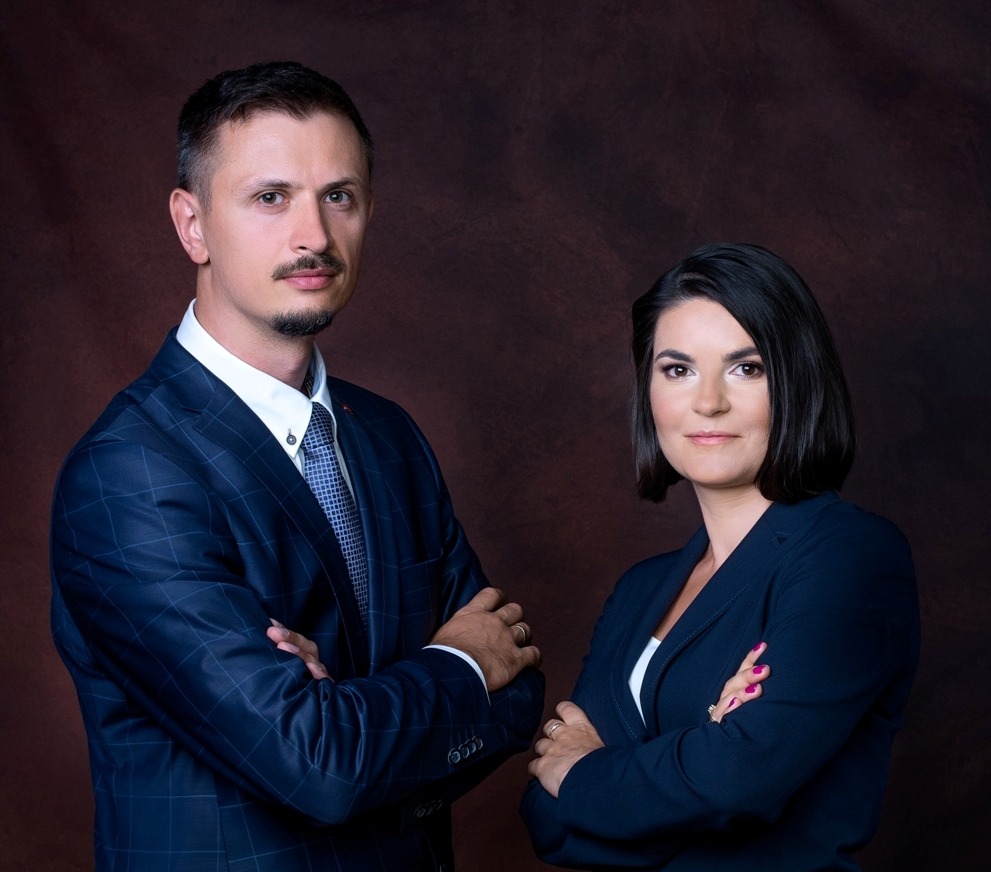 Portret dwojga prawników z Rzeszowa, mężczyzna w niebieskiej kraciastej marynarce i kobieta w granatowym garniturze, oboje z założonymi rękami na tle ciemnobrązowego tła.