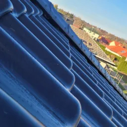 Nowe, czarne dachówki ceramiczne ułożone na dachu domu jednorodzinnego, widok pod kątem, w tle zabudowa mieszkalna i błękitne niebo.