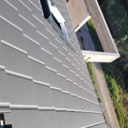 Nowy, szary dach z blachy trapezowej z oknem dachowym, widok z góry na tle niedokończonej konstrukcji domu i zieleni.