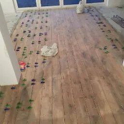 Układanie paneli podłogowych imitujących drewno z użyciem niebiesko-zielonych klipsów dystansowych, widoczny biały filar i materiały budowlane w tle.