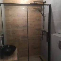 Nowoczesna kabina prysznicowa z czarnymi profilami na tle ściany wyłożonej płytkami imitującymi drewno, obok umywalka z czarną misą.
