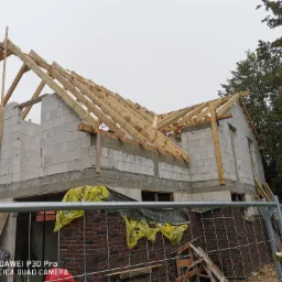 Surowy stan budowy domu z białych bloczków, widoczna konstrukcja drewniana dachu, zabezpieczenia folią i ogrodzenie tymczasowe.