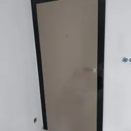 Beżowe drzwi z czarną ościeżnicą i chromowaną klamką, widoczne na tle białej ściany z wystającym gniazdkiem elektrycznym, w trakcie prac wykończeniowych.