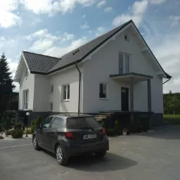 Nowoczesny dom jednorodzinny z szarą dachówką i białą elewacją, z zaparkowanym szarym samochodem Toyota Yaris na podjeździe.