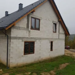 Surowy stan budynku jednorodzinnego z białych bloczków, z widocznymi brązowymi oknami i szarym dachem, na tle zielonej trawy i drzew.