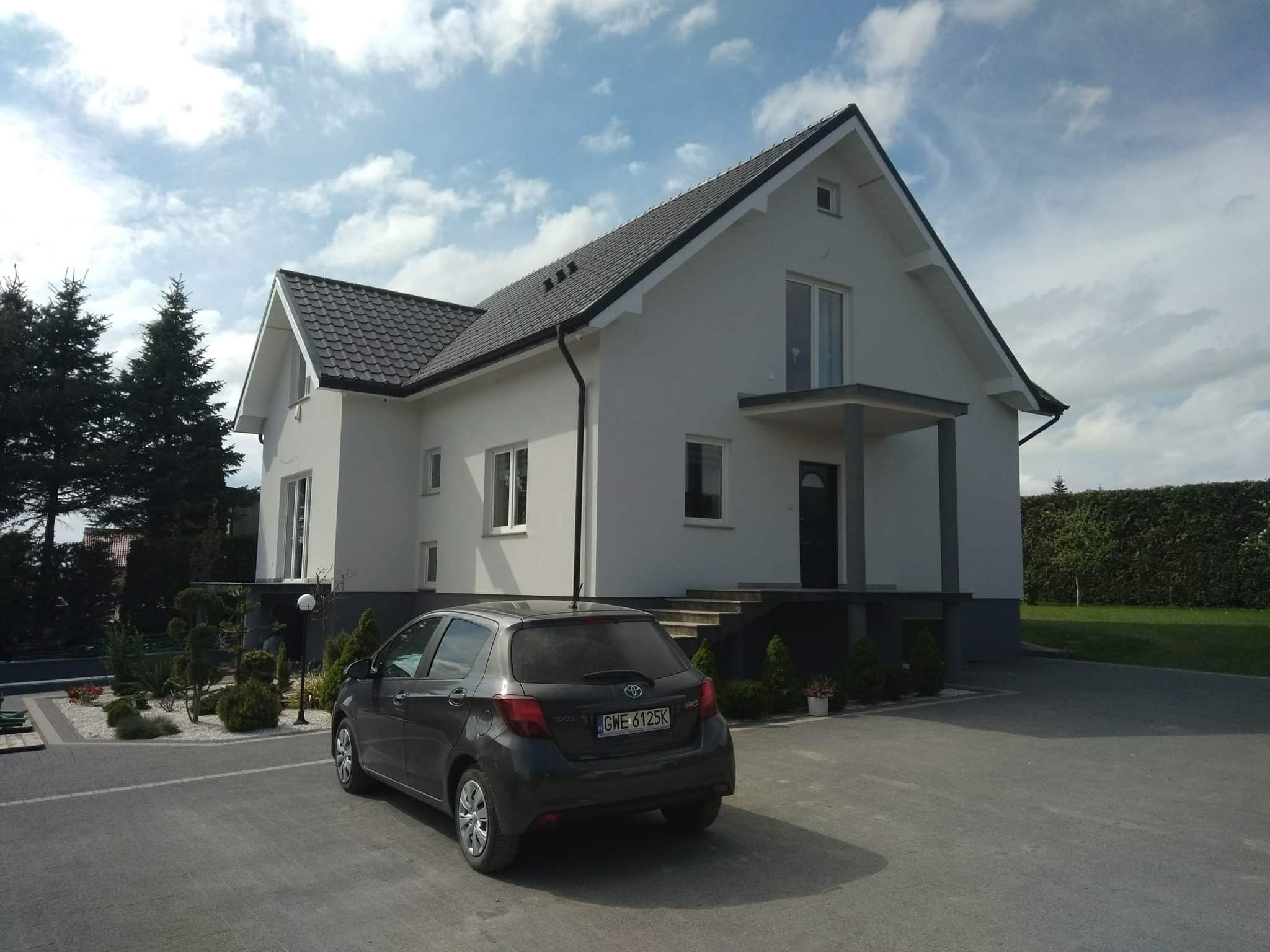 Nowoczesny dom jednorodzinny z szarą dachówką i białą elewacją, z zaparkowanym szarym samochodem Toyota Yaris na podjeździe.