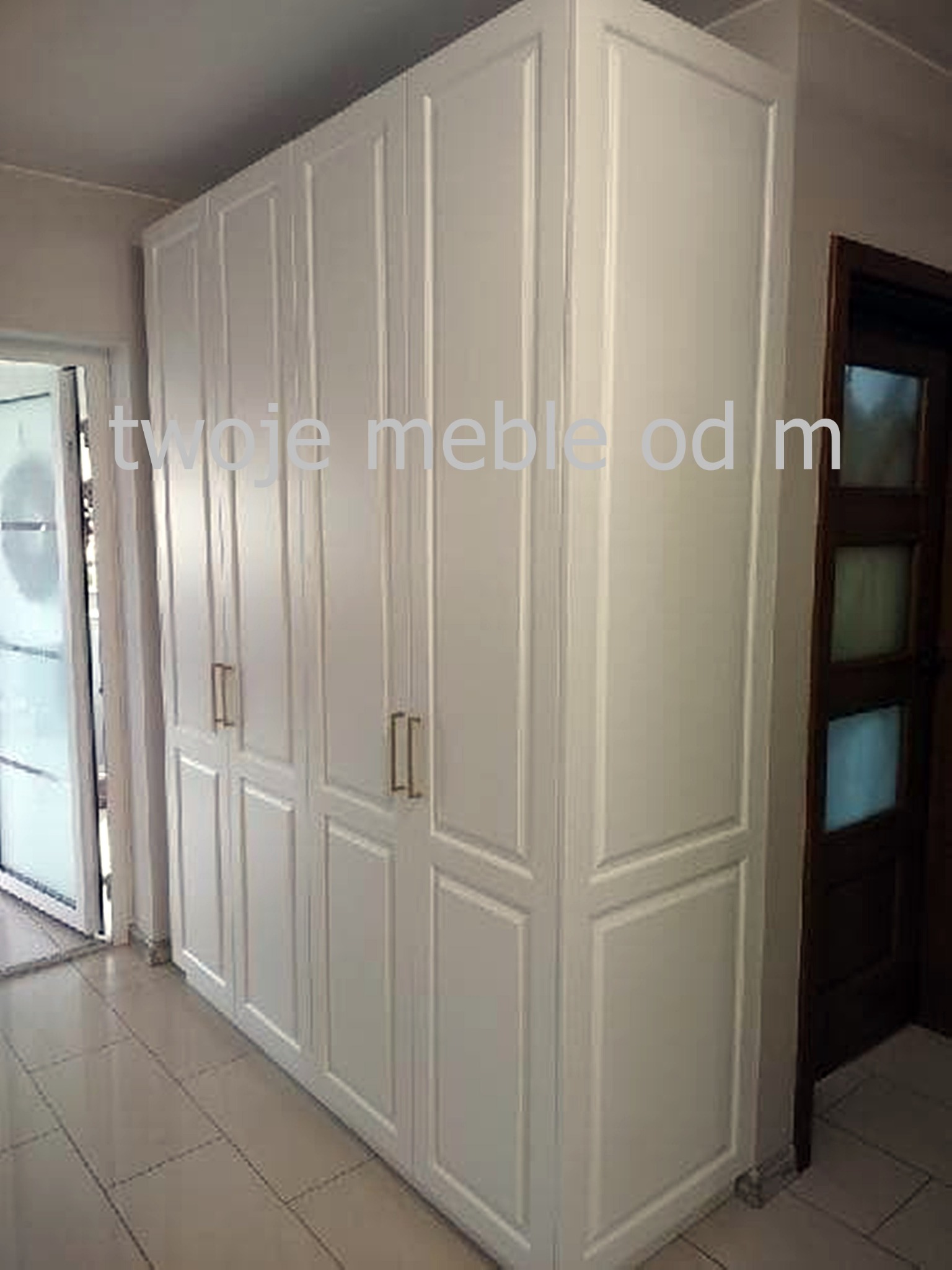 Biała, wysoka szafa z frezowanymi frontami i złotymi uchwytami, widoczna w korytarzu z płytkami podłogowymi i drzwiami po prawej stronie.