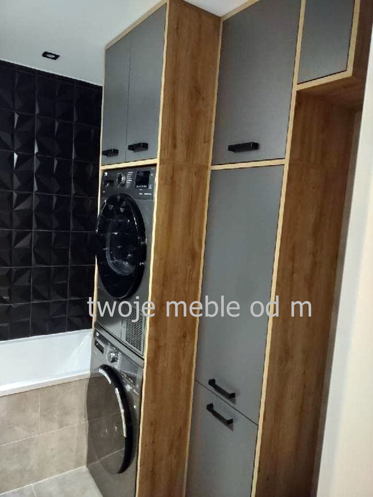 Szara szafa na wymiar z drewnianymi bokami, zabudowująca pralkę i suszarkę w łazience z czarnymi trójwymiarowymi płytkami.