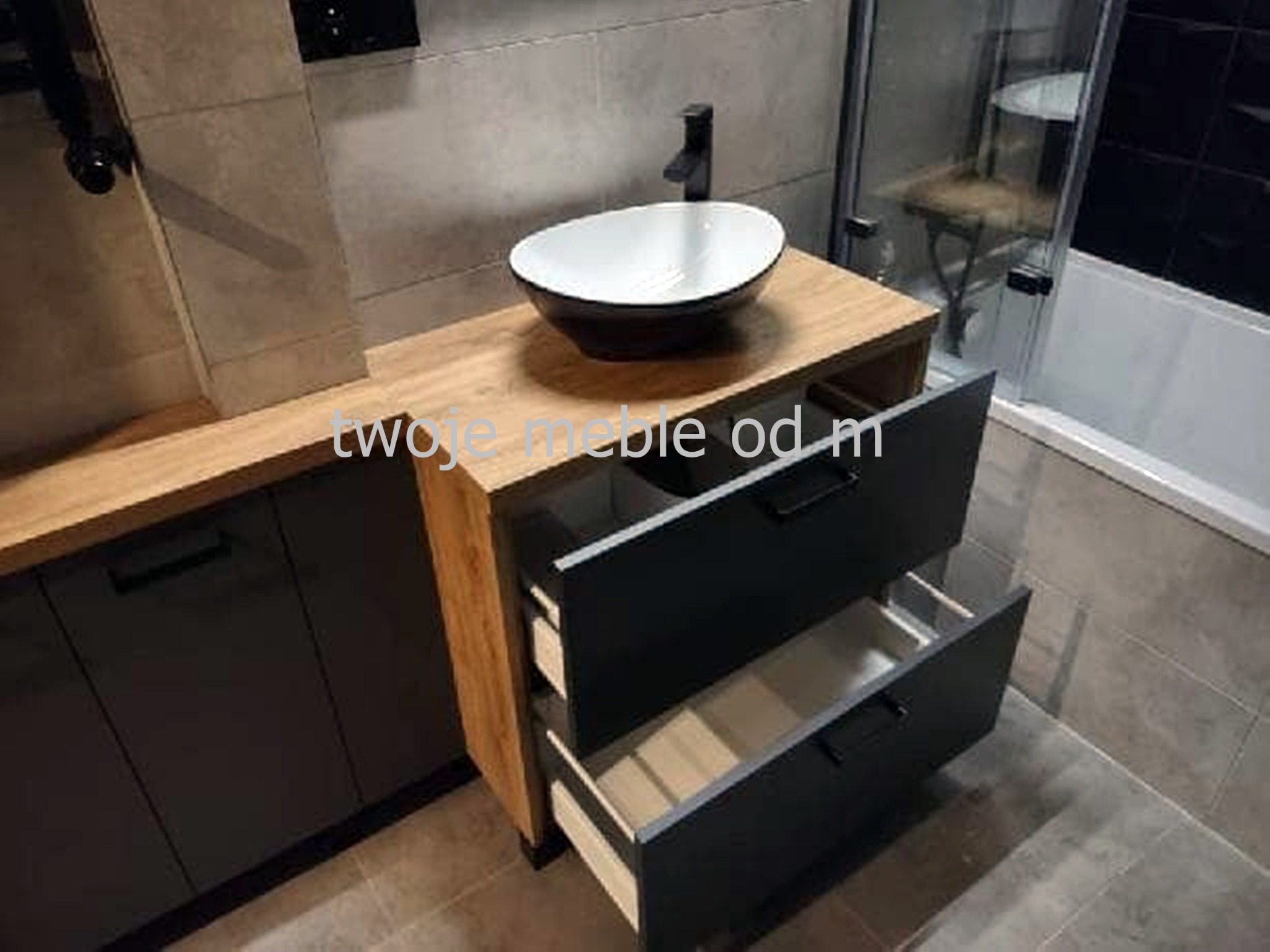 Nowoczesna łazienka z szafką pod umywalkę z blatem drewnianym i szarymi frontami, otwarte szuflady ukazują jasne wnętrze, owalna umywalka nablatowa w czarno-białej kolorystyce.