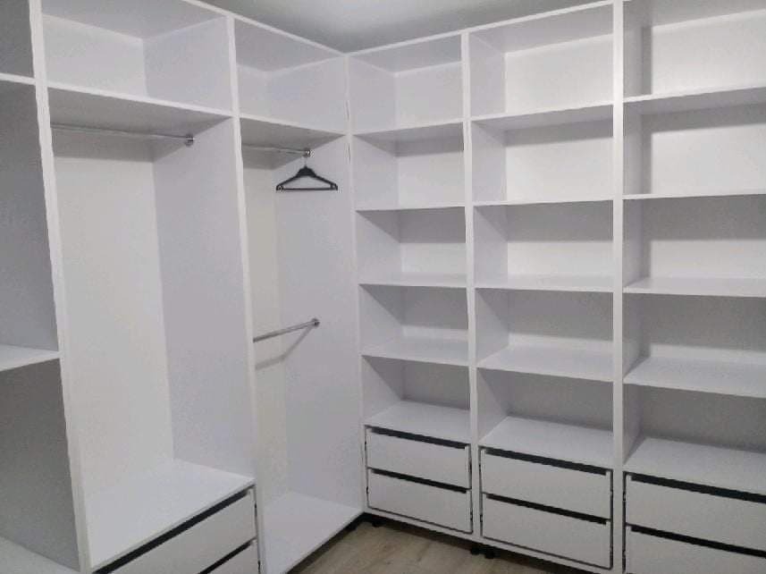 Wnętrze garderoby z białymi, modułowymi szafami i półkami, widoczny drążek na wieszaki i szuflady, jedno puste ramię wieszaka.