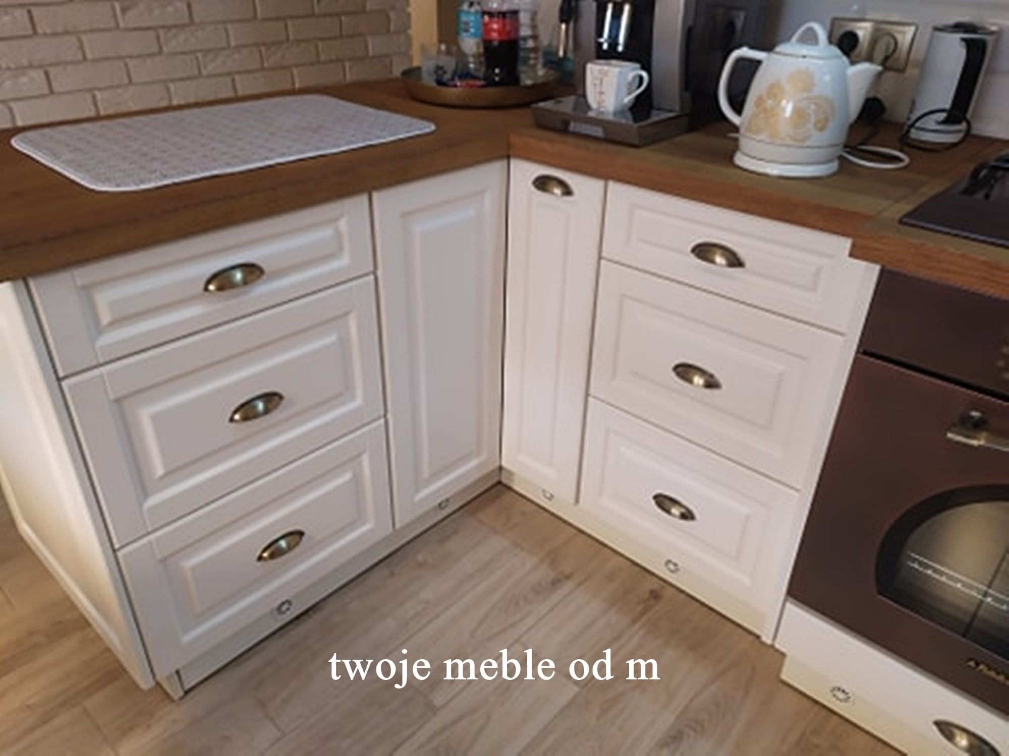 Fragment białej, narożnej zabudowy kuchennej z drewnianym blatem, widoczne szuflady z ozdobnymi uchwytami, piekarnik w brązowej obudowie i sprzęty AGD na blacie.