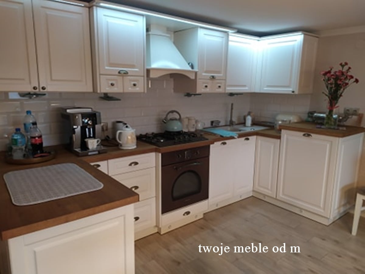 Kremowa kuchnia z drewnianym blatem, oświetleniem LED pod szafkami i sprzętem AGD, w tle biała ceglana ściana i bukiet kwiatów.