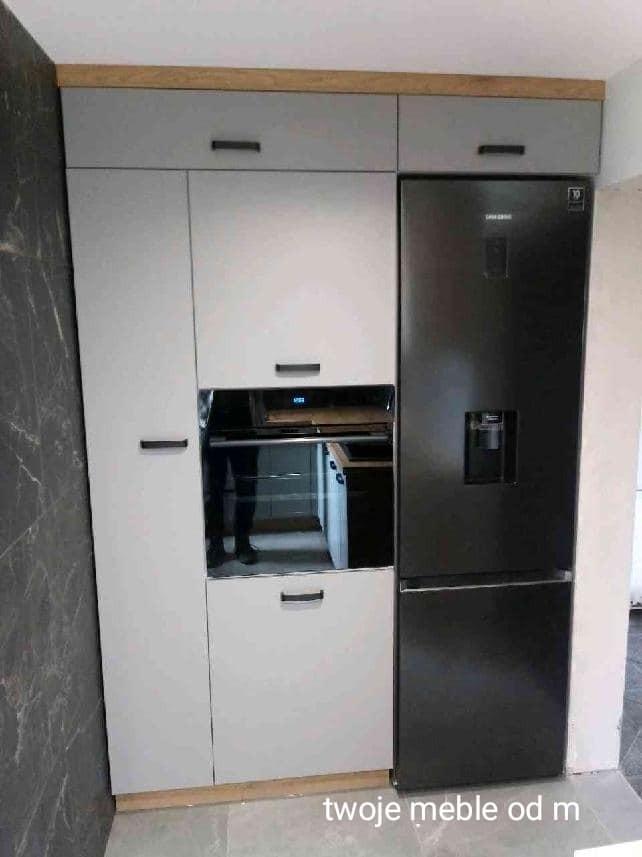 Szara zabudowa kuchenna z piekarnikiem i czarną lodówką, widoczne drewniane wykończenia górnych szafek, odbicie osoby w szklanym froncie piekarnika.