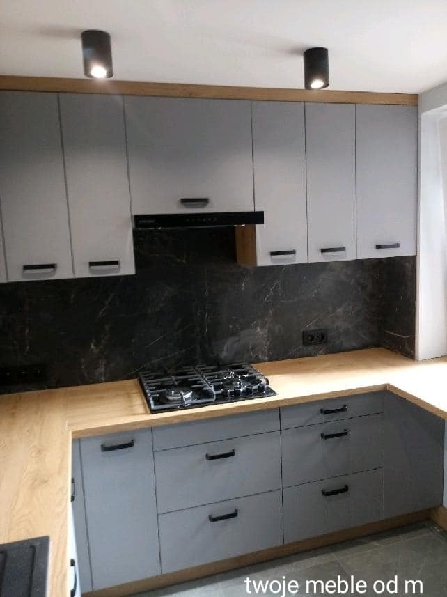 Szare szafki kuchenne z drewnianym blatem i czarnym panelem nad blatem, oświetlenie punktowe sufitowe.