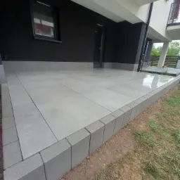 Wykończony taras z szarych płytek, obrzeża z betonowych bloczków. Nowoczesny design, czarna elewacja budynku w tle. Widok z poziomu gruntu.