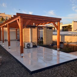 Odala Marcin Słaby - Nowoczesny taras z drewnianą pergolą i jasnymi płytkami, otoczony kamieniami. Widoczny klimatyzator i elementy zieleni. Ujęcie w ciepłym świetle zachodzącego słońca.