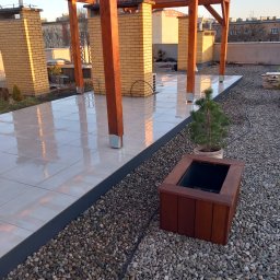 Odala Marcin Słaby - Wykończony taras na dachu w Rzeplinie z drewnianą pergolą, jasnymi płytkami i kamiennym otoczeniem. Widoczna donica z drzewkiem.