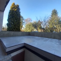 Odala Marcin Słaby - Wykończony taras z szarych płyt, otoczony niską ścianką. Widok na drzewa i błękitne niebo. Schody prowadzą w dół. Minimalistyczny design.