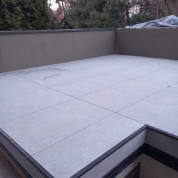 Odala Marcin Słaby - Wykończony taras z jasnoszarych płyt, obrzeża z ciemnego tworzywa. Widoczne drzewa w tle. Minimalistyczny design, świeżo po realizacji.