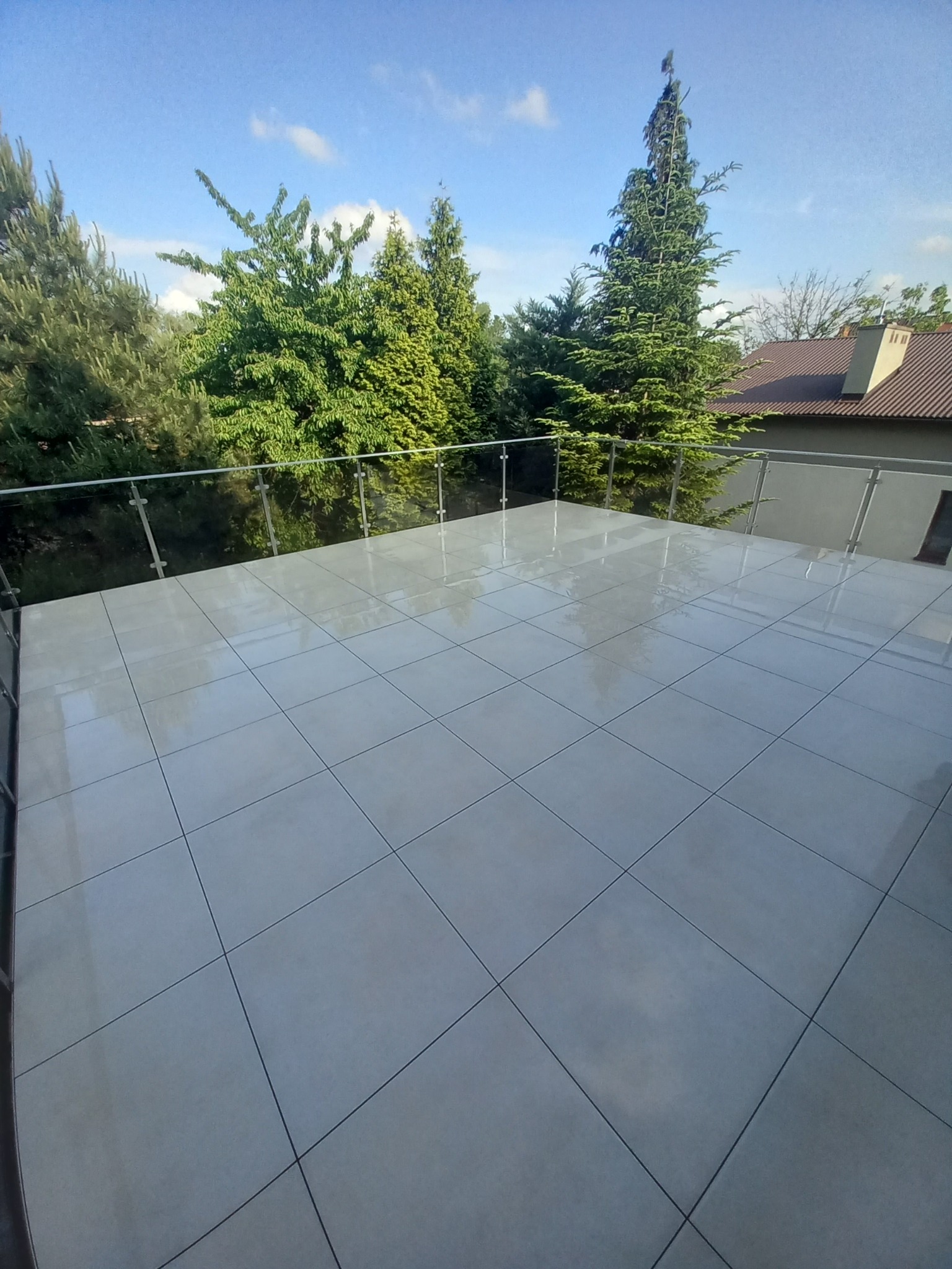 Błyszczące, jasnoszare płytki na balkonie z szklaną balustradą, widok na zieleń drzew i fragment domu w tle, słoneczny dzień.