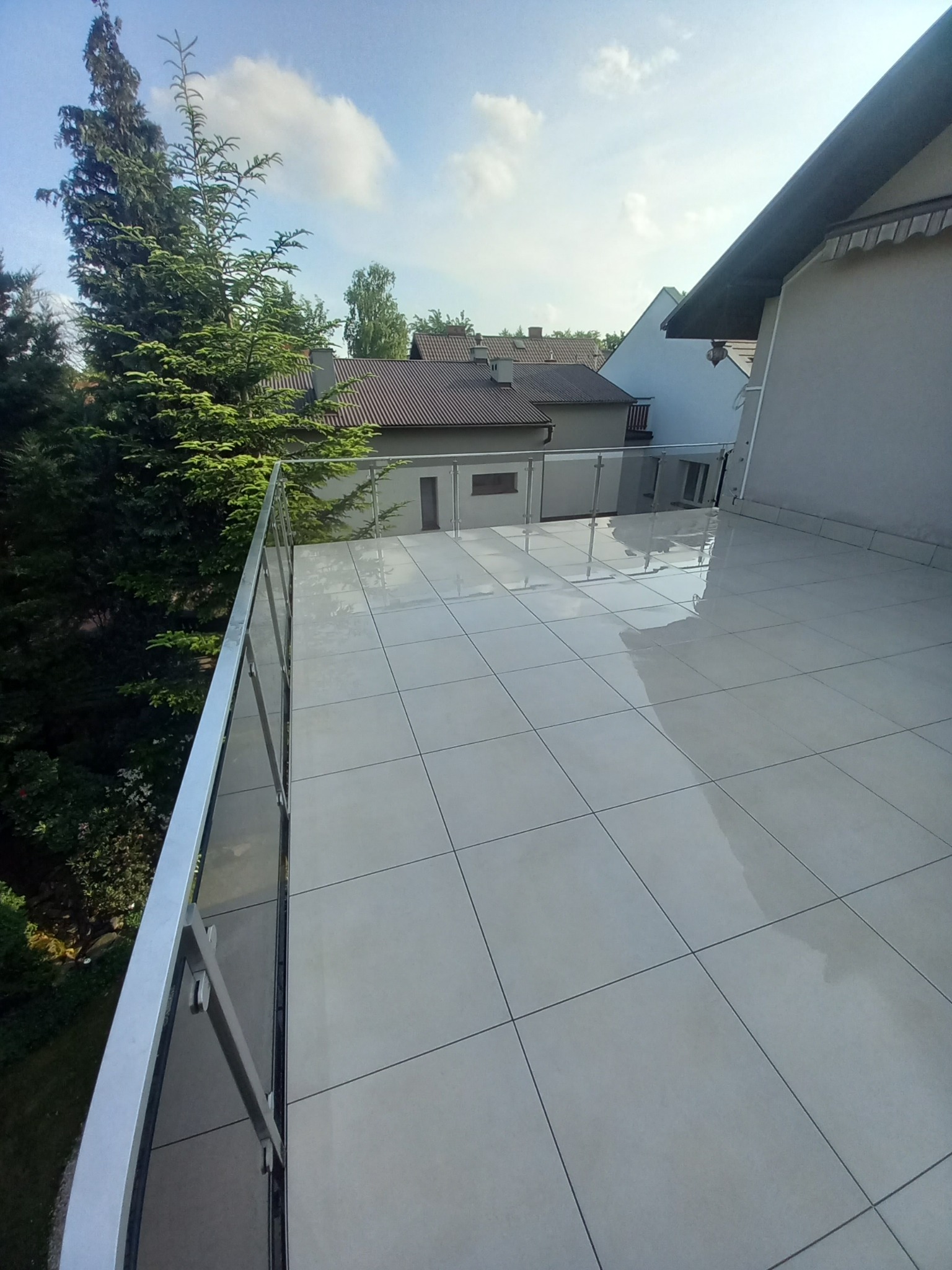Błyszcząca, jasna podłoga balkonu z szarych płytek, otoczona nowoczesną balustradą ze stali i szkła w Rzeplinie. Widok na zieleń i budynki.