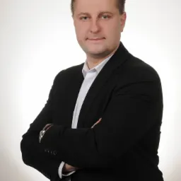 Portret mężczyzny w czarnym garniturze i białej koszuli, z rękami skrzyżowanymi na piersiach, patrzącego w obiektyw na jasnym, jednolitym tle.