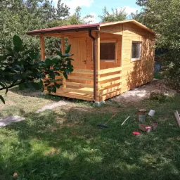 Świeżo wybudowana, surowa, drewniana konstrukcja z czerwonym dachem, prawdopodobnie domek narzędziowy lub mała altana, na trawniku widoczne narzędzia i puszki po farbach.