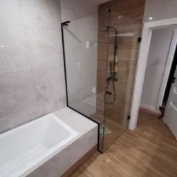 Nowoczesna łazienka z kabiną prysznicową typu walk-in, obudowaną wanną i drewnopodobnymi płytkami podłogowymi oraz szarymi płytkami na ścianach.