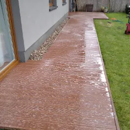 Brązowy taras z desek kompozytowych, świeżo umyty, przylegający do jasnoszarego budynku w Józefowie. Widoczne kamienie ozdobne wzdłuż ściany oraz zielony trawnik.