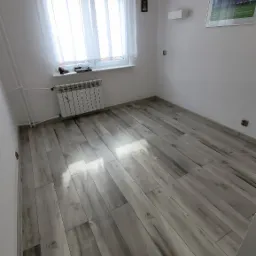 Pokój z szarymi panelami podłogowymi imitującymi drewno, oświetlony lampą w kształcie piłki nożnej, z grzejnikiem pod oknem i dekoracjami o tematyce piłkarskiej.