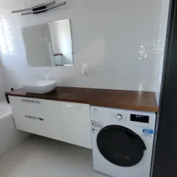 Biała łazienka z umywalką nablatową, szafką z szufladami pod drewnianym blatem i pralką Beko na tle białych płytek z subtelnym wzorem.