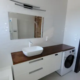 Nowoczesna łazienka w Karolinkach z białą, owalną umywalką nablatową na drewnianym blacie, podwieszane szafki z szufladami i pralka obok, całość w jasnej tonacji z dekoracyjnym oświetleniem...