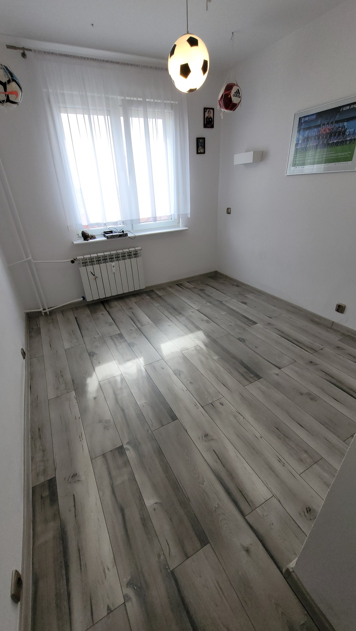 Pokój z szarymi panelami podłogowymi imitującymi drewno, oświetlony lampą w kształcie piłki nożnej, z grzejnikiem pod oknem i dekoracjami o tematyce piłkarskiej.