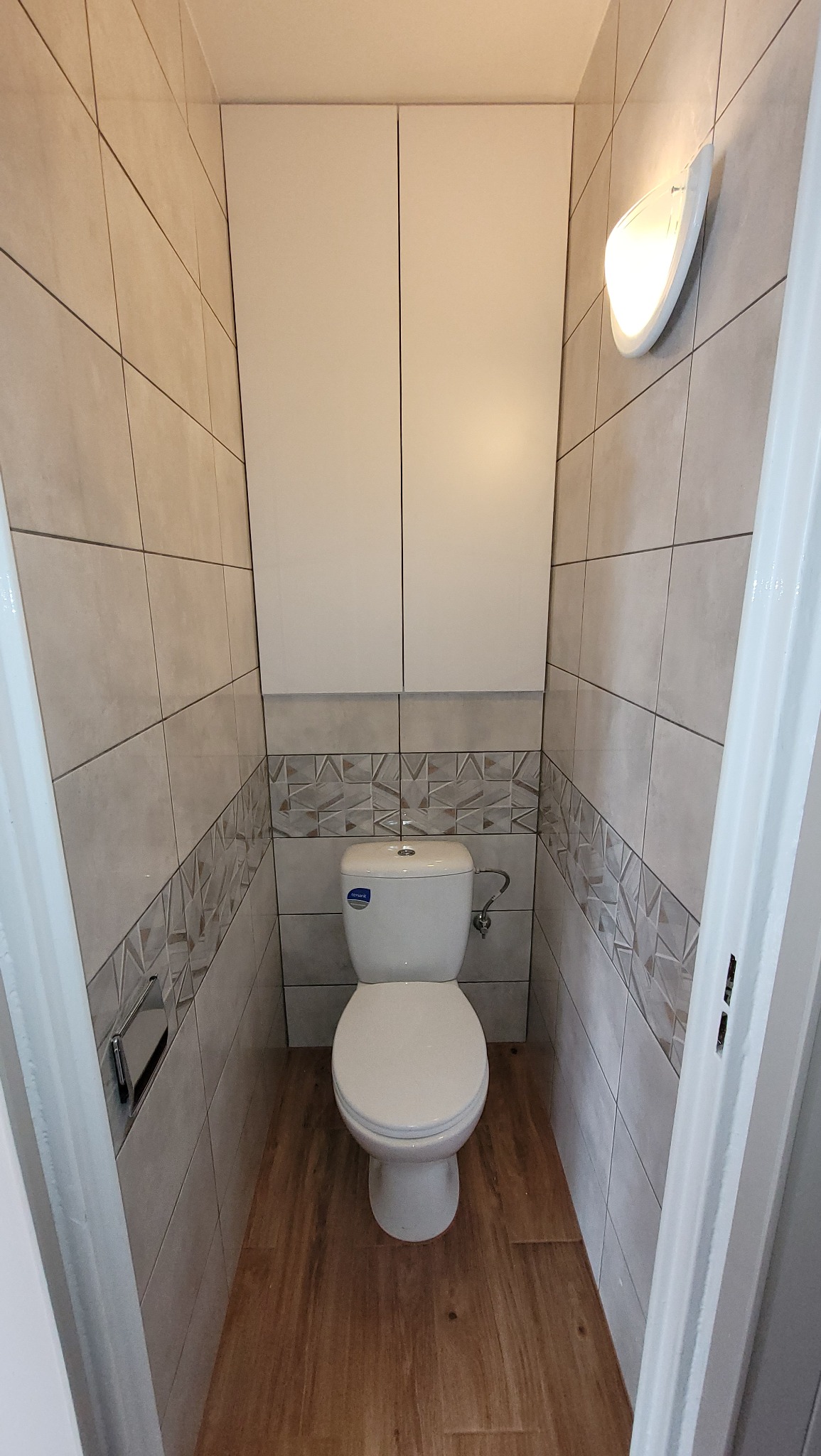 Mała toaleta z białym kompaktem WC, zabudową szafek nad nim, wykończona płytkami imitującymi drewno na podłodze i szarymi płytkami z dekoracyjnym pasem na ścianach.