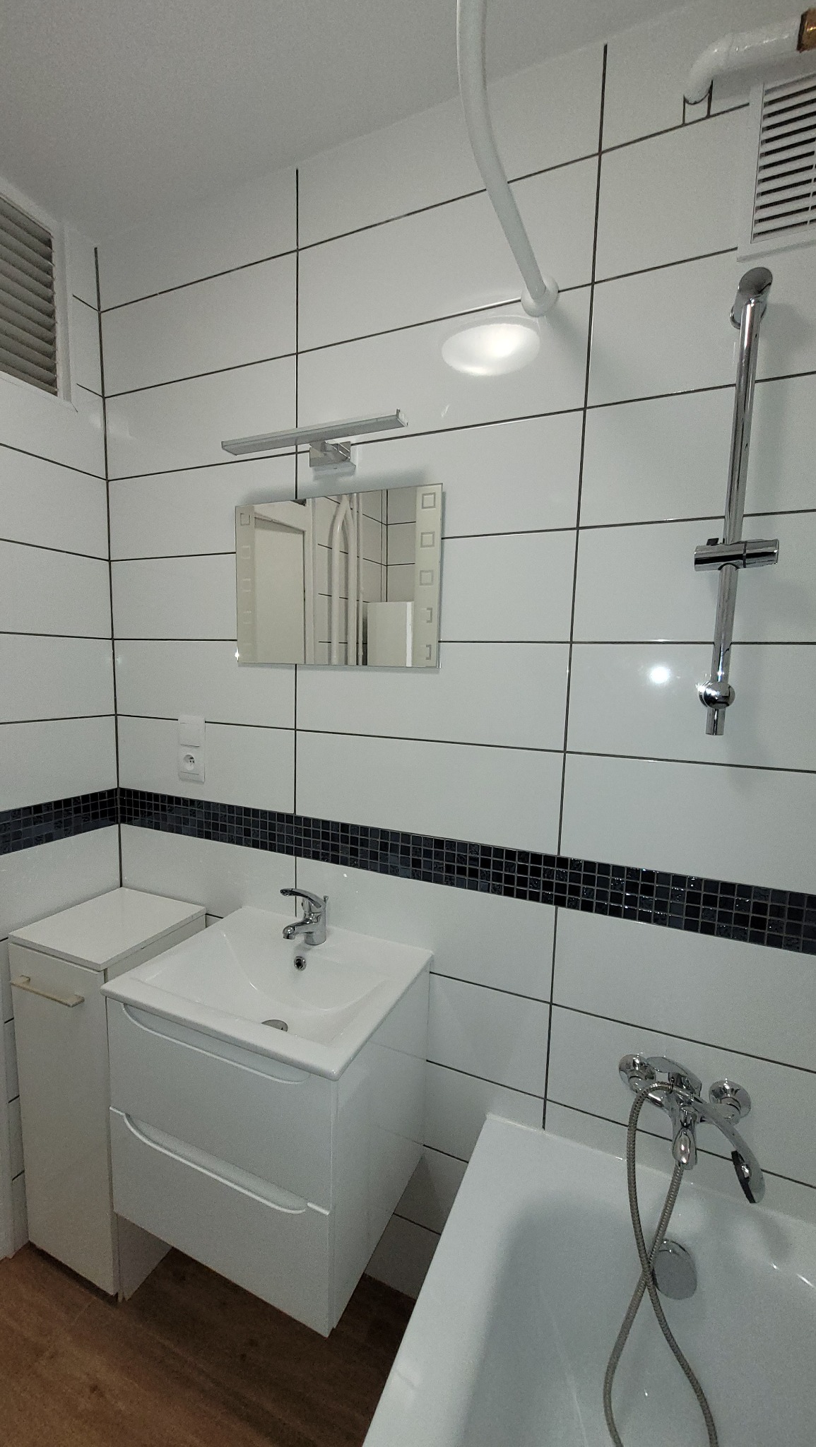 Biała łazienka z umywalką nablatową, lustrem z podświetleniem LED, wanną i kabiną prysznicową z chromowaną armaturą, wykończona białymi płytkami z czarną mozaiką.