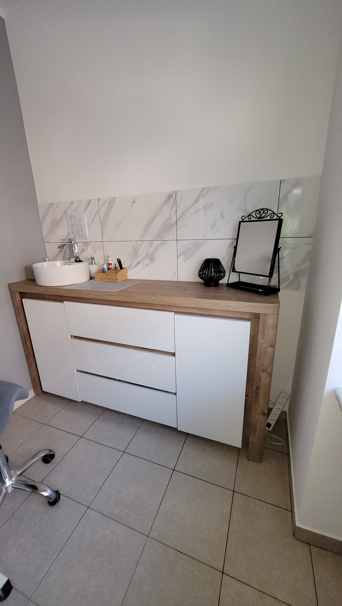 Nowoczesna łazienka z białą umywalką nablatową, szafką z frontami w kolorze białym i blatem imitującym drewno, wykończona płytkami imitującymi marmur. Na blacie dekoracje: czarna ażurowa kula...