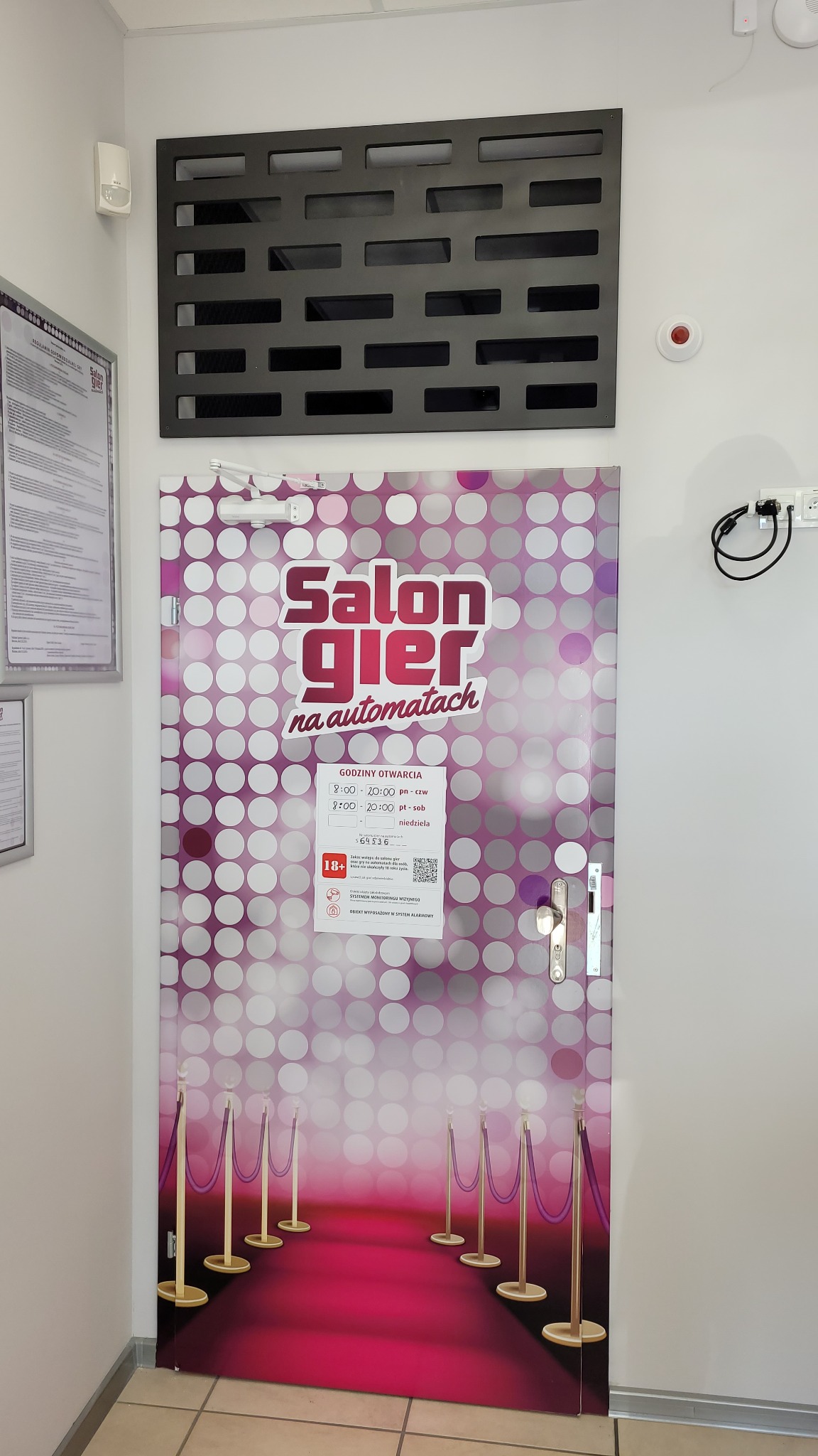 Drzwi oklejone folią z grafiką salonu gier na automatach, z motywem czerwonego dywanu i godzinami otwarcia.