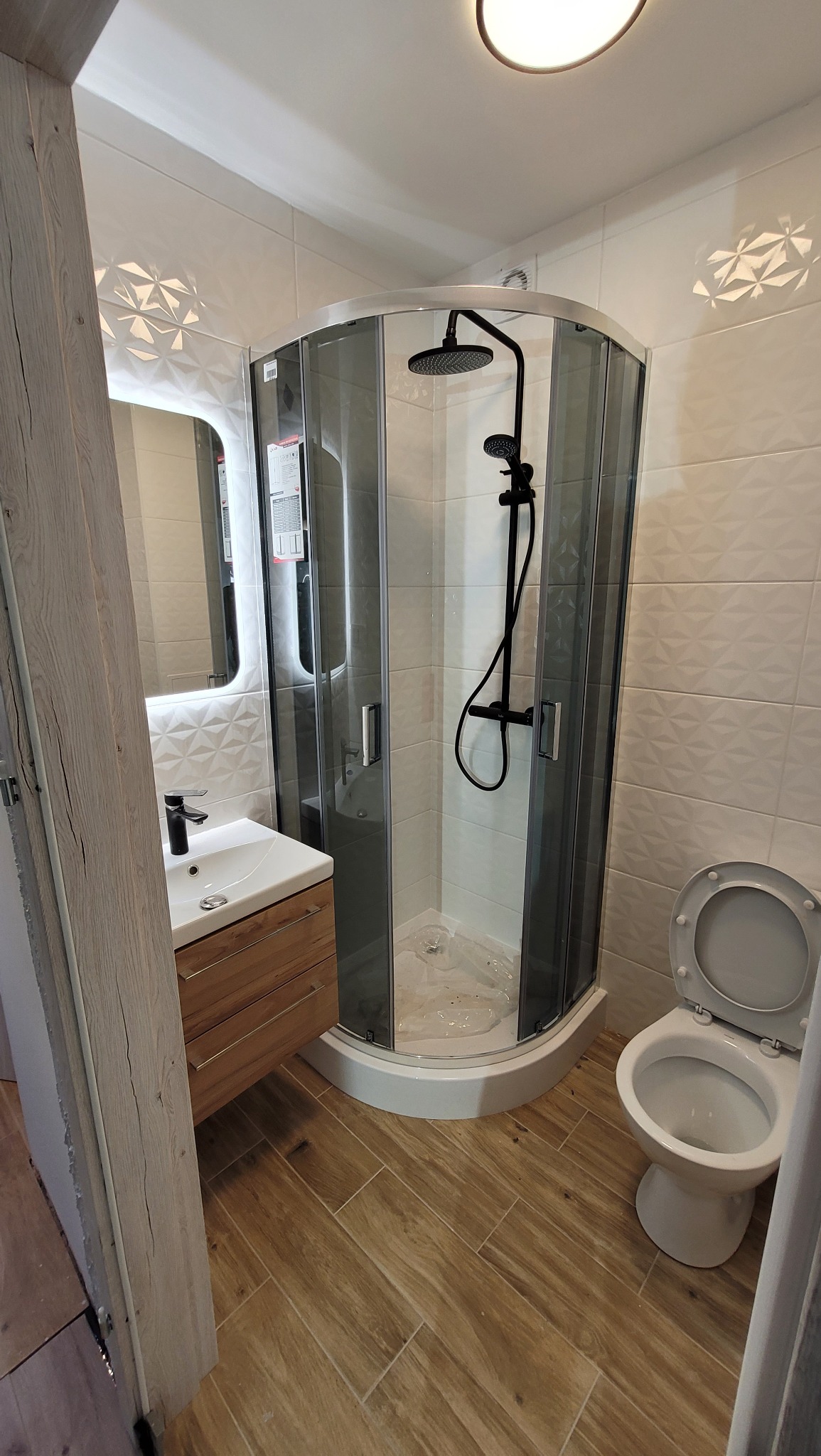 Nowoczesna łazienka z kabiną prysznicową z czarną armaturą, drewnianą szafką pod umywalką i toaletą z otwartą klapą, wykończona białymi płytkami z geometrycznym wzorem i drewnopodobną podłogą.