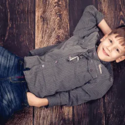 Uśmiechnięty chłopiec w dżinsach i koszuli w paski leży na ciemnym drewnianym tle, z okularami na koszuli.