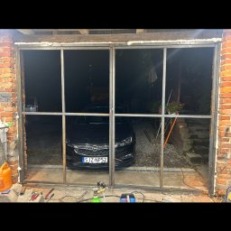 Usługi spawalnicze Bachlaj Wojciech - Surowa, metalowa konstrukcja ramy okiennej w murze z czerwonej cegły. W tle widoczny samochód i narzędzia ślusarskie. Etap prac montażowych.