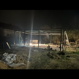 Usługi spawalnicze Bachlaj Wojciech - Nocna budowa metalowej konstrukcji z widocznym spawaczem i iskrzącym spawem. W tle dom i drzewa. Konstrukcja w trakcie montażu, widoczne elementy fundamentów.