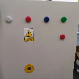 Szara szafa sterownicza z kolorowymi przyciskami, ostrzeżeniem o napięciu elektrycznym i etykietą 'Wyłącznik Główny', częściowo zasłonięta ręką z czarną opaską fitness.