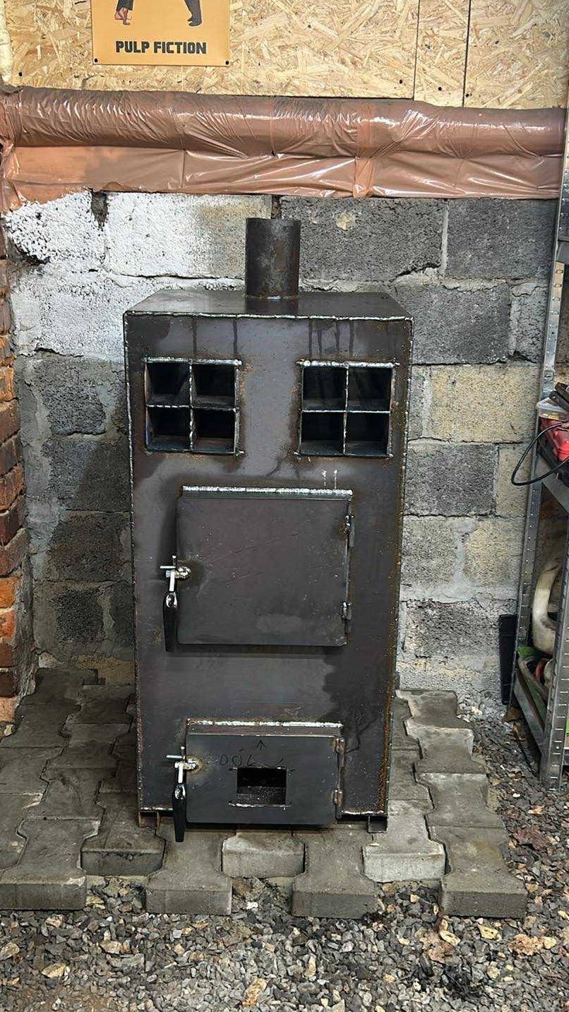 Metalowy piec z dwoma małymi okienkami i dwoma drzwiczkami, ustawiony na betonowej kostce brukowej, na tle ściany z betonowych pustaków. Nad piecem plakat Pulp Fiction.