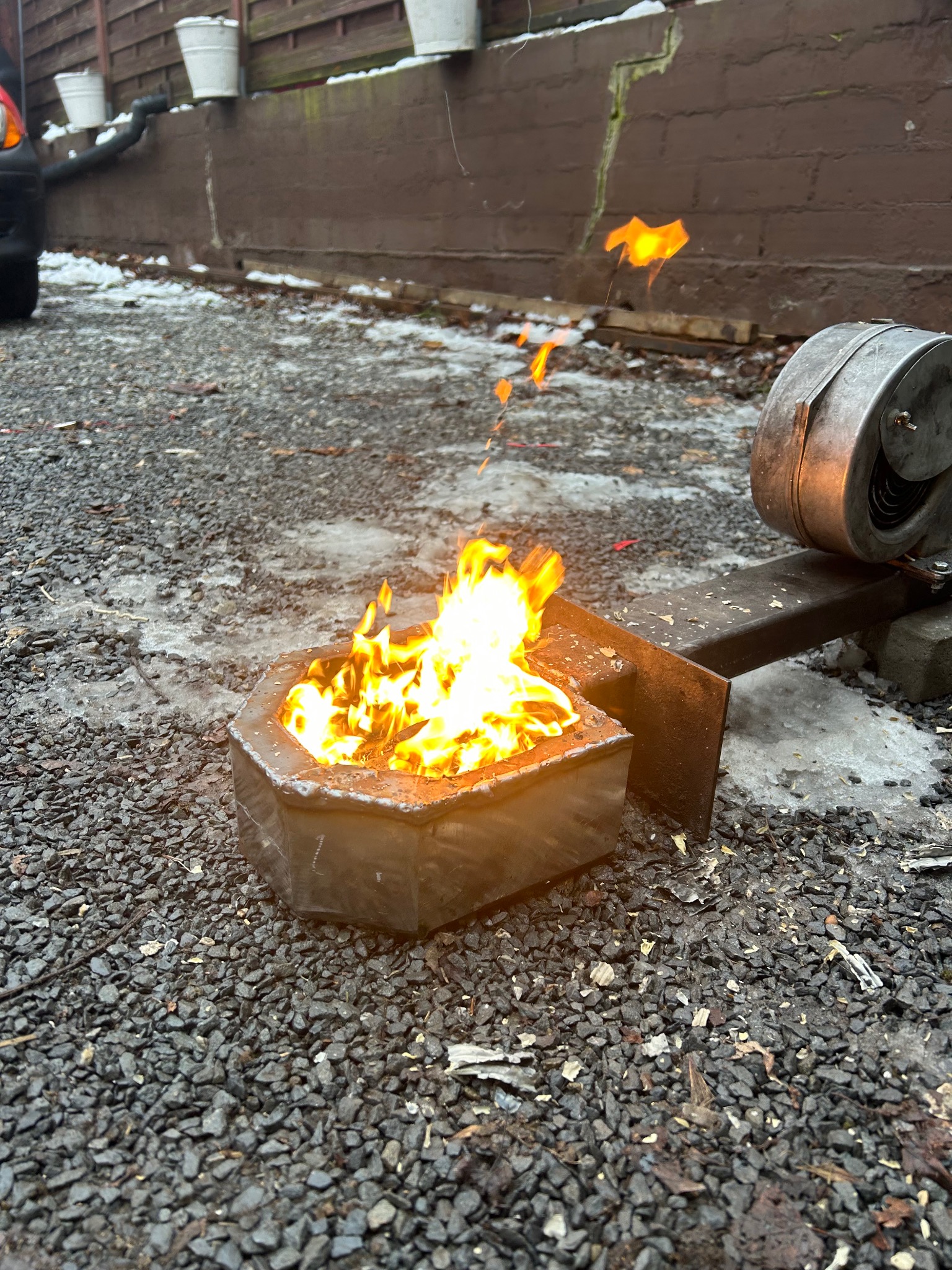 Ośmiokątny, metalowy palenisko z płomieniami na żwirowym podłożu, z fragmentami śniegu. W tle brązowy mur i fragment samochodu. Nietypowe ujęcie paleniska.