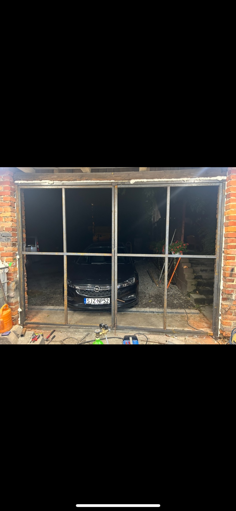 Surowa, metalowa konstrukcja ramy okiennej w murze z czerwonej cegły. W tle widoczny samochód i narzędzia ślusarskie. Etap prac montażowych.