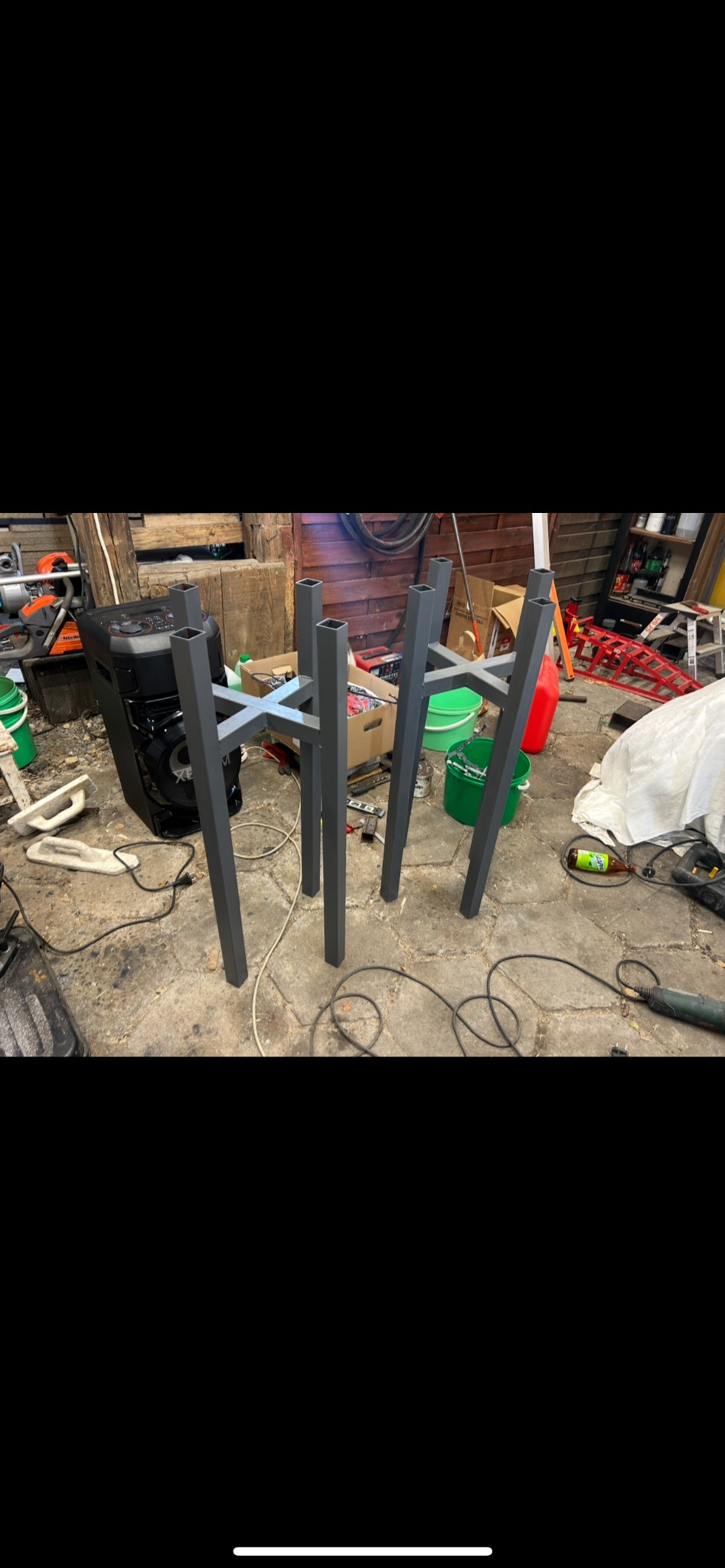 Metalowa konstrukcja stołu z profili kwadratowych, spawana krzyżowo, w szarym kolorze, na tle warsztatu z narzędziami i sprzętem.