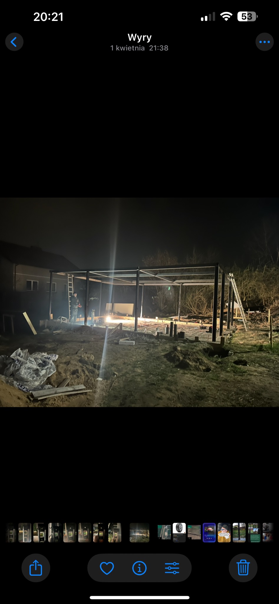 Nocna budowa metalowej konstrukcji z widocznym spawaczem i iskrzącym spawem. W tle dom i drzewa. Konstrukcja w trakcie montażu, widoczne elementy fundamentów.