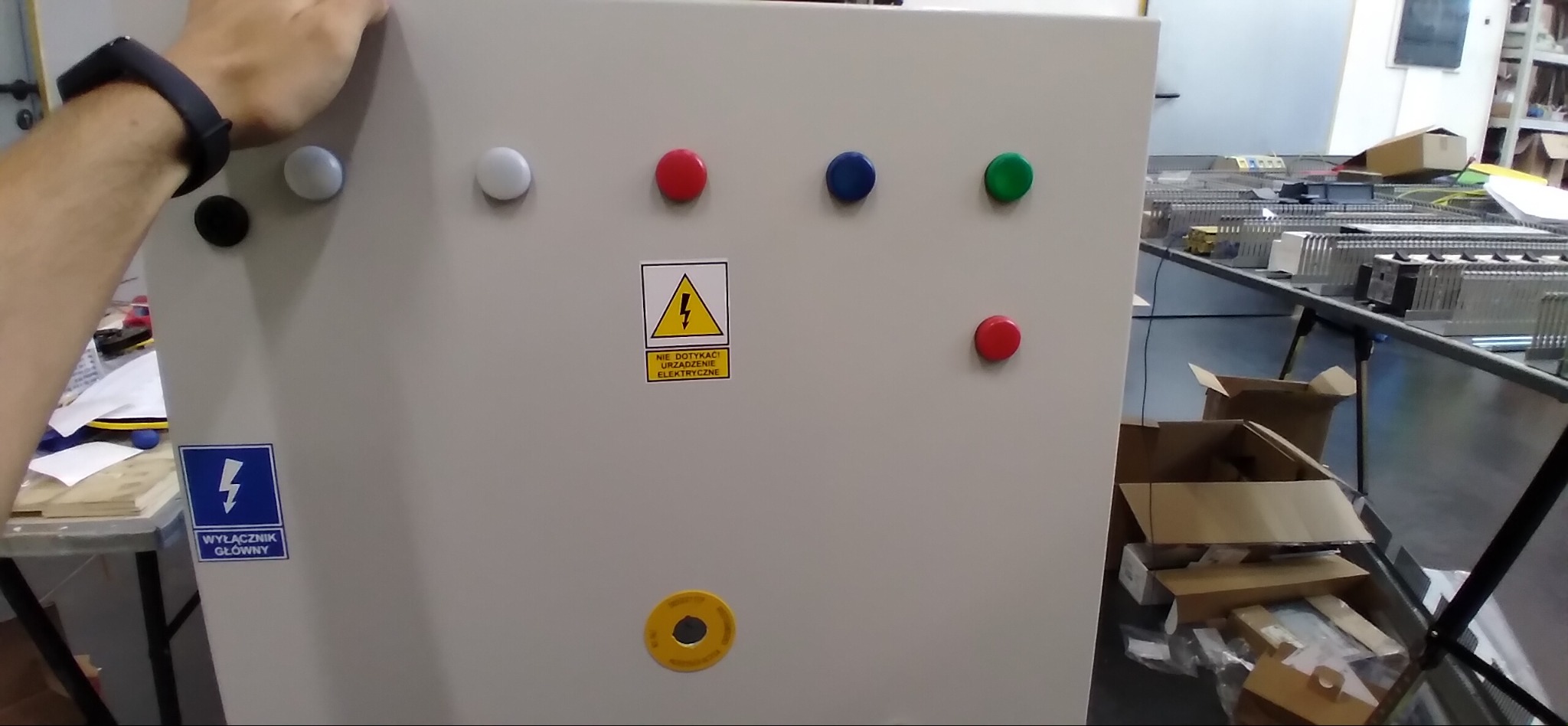 Szara szafa sterownicza z kolorowymi przyciskami, ostrzeżeniem o napięciu elektrycznym i etykietą 'Wyłącznik Główny', częściowo zasłonięta ręką z czarną opaską fitness.