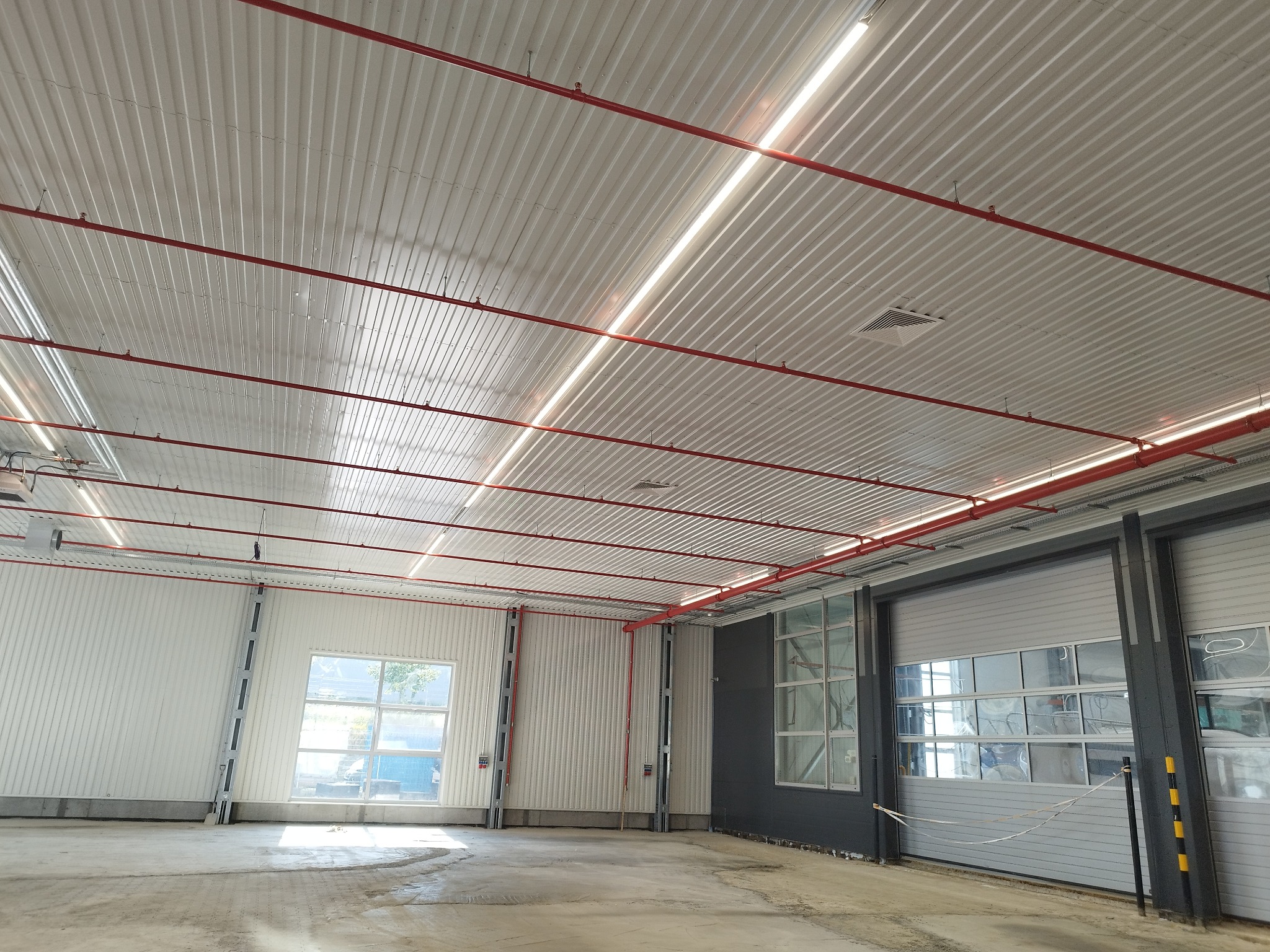 Wnętrze hali przemysłowej z widocznymi instalacjami pod stropem: czerwone rury przeciwpożarowe, białe panele oświetleniowe LED, oraz instalacje wentylacyjne.